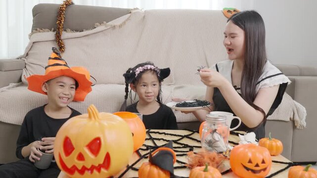 Asian family enjoying Halloween fun at home, children wearing costumes, playing with toy spiders, pumpkins, webs decorate table, festive atmosphere for happy family celebration.