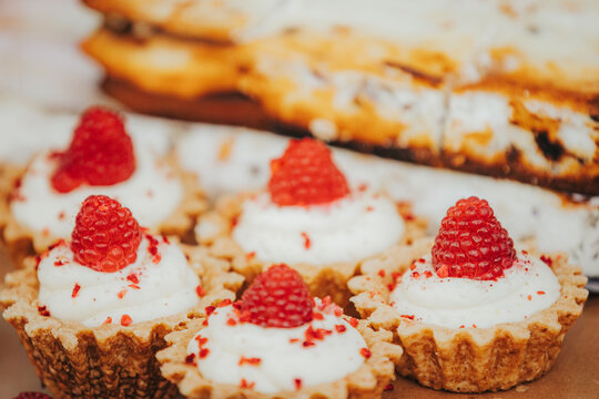 Delicious mini tart desserts with creamy frosting topped with fresh raspberries arranged on a wooden surface with additional baked goods in the background