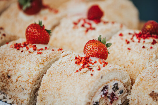 Delicious rolled cake dessert topped with fresh strawberries and red sprinkles, displayed on a white plate against a blurred background of sweet treats