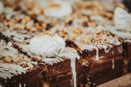 Chocolate cake topped with meringue and crushed nuts, drizzled with white chocolate, displayed on a wooden surface in a bakery setting
