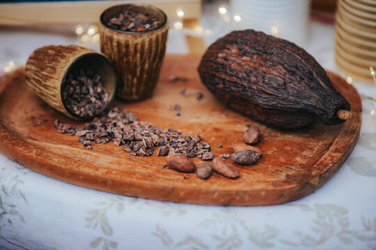 Wooden platter with cacao pods, cocoa nibs, and two decorative bowls filled with chocolate pieces on a textured tablecloth with soft lighting