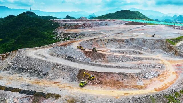 Aerial view of a terraced open pit quarry with excavators and haul trucks, surrounded by forested hills under bright, diffuse overcast light.