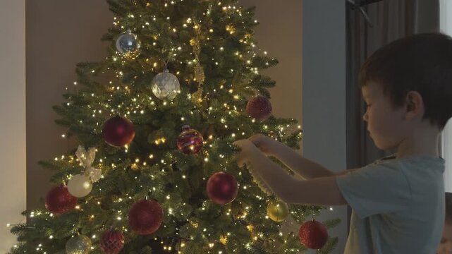 Child carefully hanging red ornament on Christmas tree. Cozy festive home atmosphere. Perfect for holiday marketing, parenting content and Christmas campaigns.