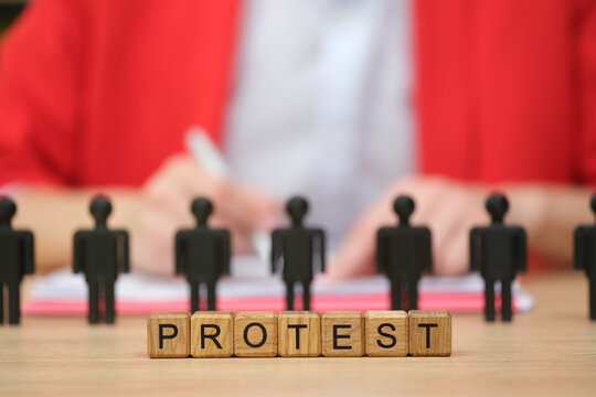 Small black figures stand in line, highlighting the word protest made from wooden blocks. A person in the background takes notes, capturing the essence of activism and community spirit.