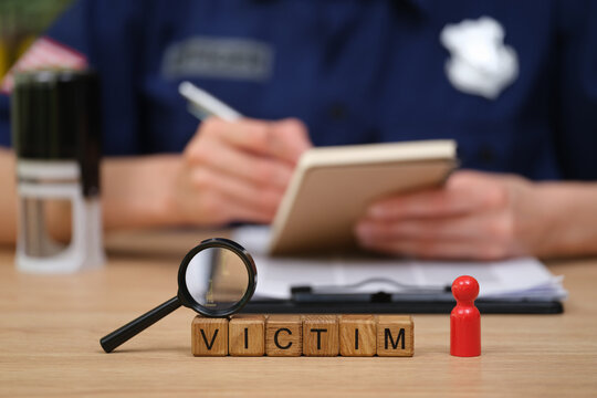Investigation scene set on a wooden table features a magnifying glass and colorful game pieces, with a person in a police uniform jotting notes. The word VICTIM is clearly spelled out.