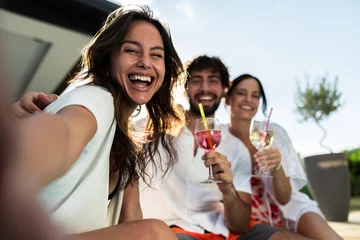 Three friends smile and pose for a selfie while enjoying refreshing cocktails in a vibrant outdoor setting. The sun shines brightly, creating a joyful and relaxed summer atmosphere with greenery in th © milanmarkovic78