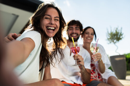 Three friends smile and pose for a selfie while enjoying refreshing cocktails in a vibrant outdoor setting. The sun shines brightly, creating a joyful and relaxed summer atmosphere with greenery in th