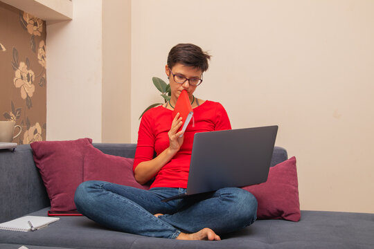 Person sitting on couch at home using laptop and holding a notebook while deep in thought