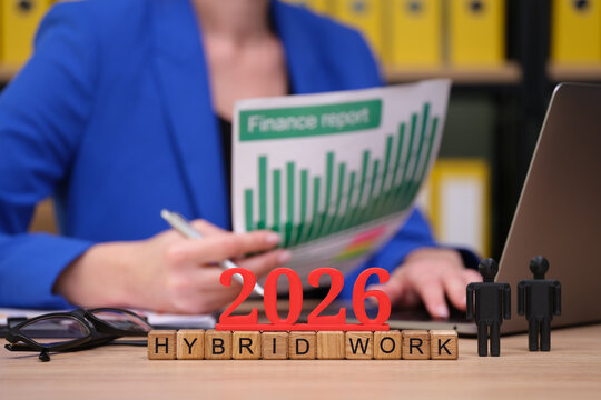 A person in a blue blazer reviews a finance report in an office setting. Wooden blocks spell out 2026 and Hybrid Work, highlighting the shift in workplace dynamics and trends.