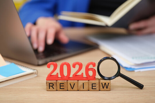 In a bright workspace, a pair of hands holds a book while a laptop rests nearby. The year 2026 stands boldly in red, symbolizing an important review moment.