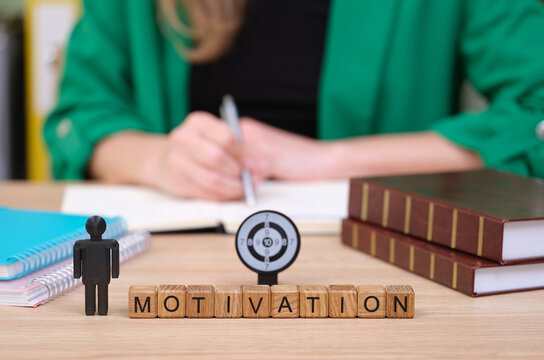 A small figure stands next to wooden blocks spelling 'MOTIVATION', while a woman in a green jacket writes in a notebook at her workspace, books stacked nearby, creating a productive atmosphere.