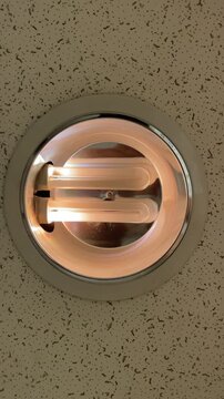 centered top-down view of recessed ceiling downlight containing two illuminated u-shaped fluorescent tubes inside round chrome reflector and white trim, mounted in
