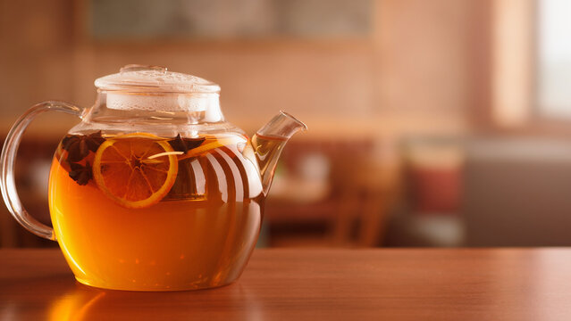 Glass teapot with spiced herbal tea and dried orange slices with blurred tea lounge background banner.