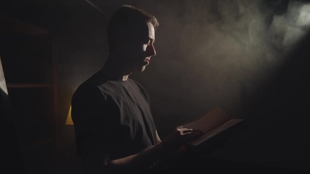 Young man is reading a book in a dimly lit room. The light from the lamp casts a warm glow on the pages of the book, cinematic scene