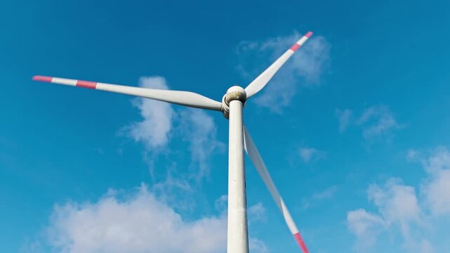 A white wind turbine with red blade markings stands against a vivid blue sky, with scattered white clouds illuminated by bright sunlight.