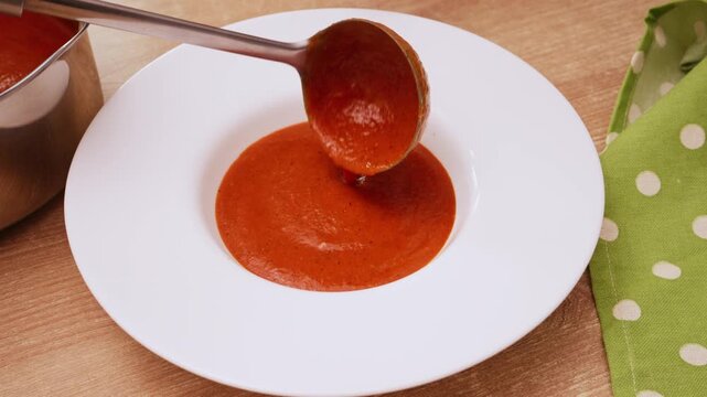 Hot tomato and red pepper soup being poured from ladle into white ceramic bowl. Fresh steam rising from rich homemade vegetable soup serving.