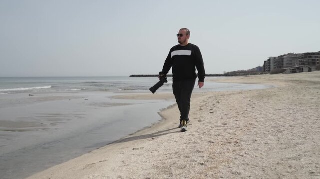 Photographer is holding a camera with a telephoto lens walking on a sandy beach
