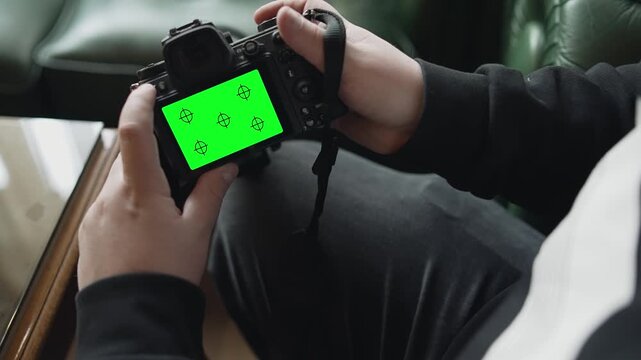 A man is holding a camera with a green screen. He is sitting on a chair
