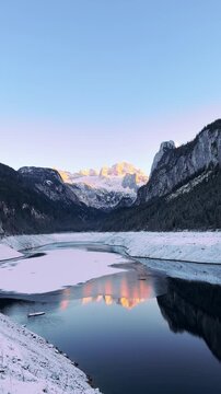 Cinematic sunset over Alpine lake in winter mountains. Winter mountain landscape. Golden hour reflection in calm mountain lake. Alpine peaks illuminated by orange light of setting sun.