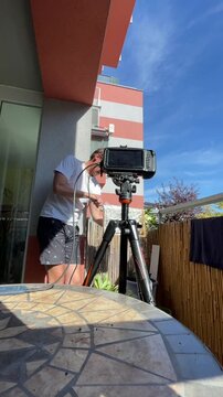 man on balcony leaning forward to connect cables and adjust controls on tripod-mounted monitor, focused hands-on setup suggesting equipment testing or home, with