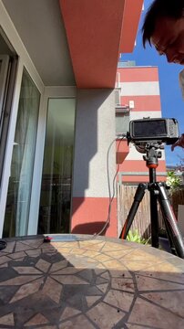 person adjusts tripod-mounted monitor while standing on small apartment balcony under bright midday sun, hands and body showing active setup and measurement near