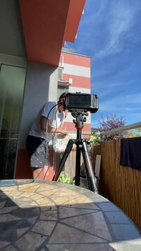 man in white t-shirt and dark shorts adjusts clear foil-wrapped container near balcony railing while standing behind monitor mounted on tripod, vertical low-angle