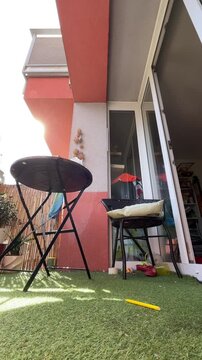 low-angle view of small urban balcony showing round folding metal table and woven rattan chair with pale cushion beside open sliding glass door