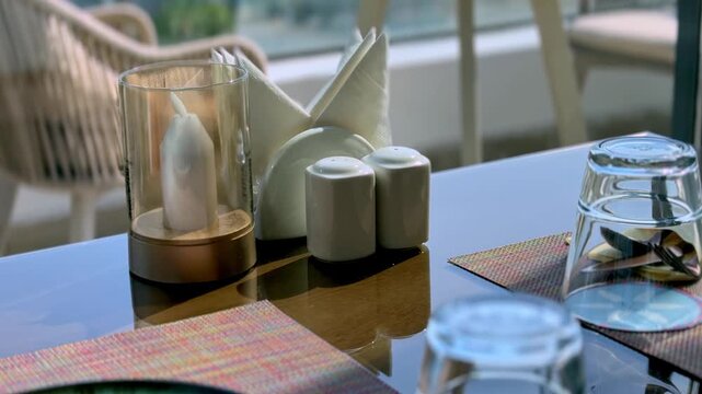 Close-up static shot of an empty outdoor restaurant table setting in sunny daylight. The glass table features a candle, salt shakers, and empty glasses with blurred woven chairs in the background.