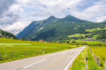 Obraz premium A winding alpine road stretches through green fields with mountains in the background. Houses dot the landscape under a cloudy sky. The area is vibrant with nature's colors.