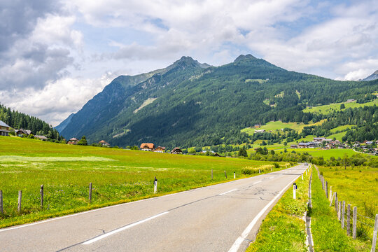 A winding alpine road stretches through green fields with mountains in the background. Houses dot the landscape under a cloudy sky. The area is vibrant with nature's colors.