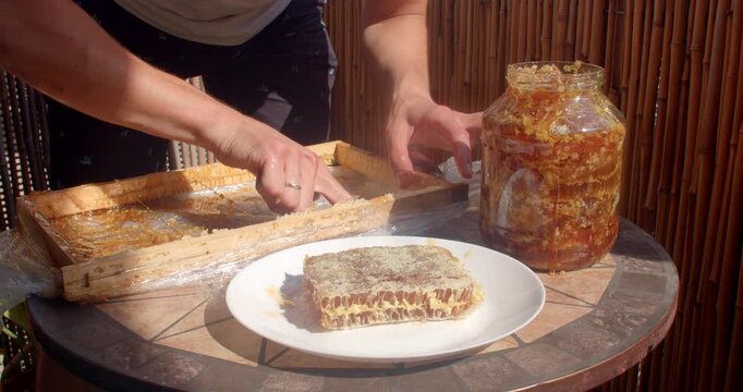closeup of hands pressing honeycomb comb, bringing sticky fingers toward mouth, tasting honey beside large jar on sunlit outdoor patio table with warm shadows amber