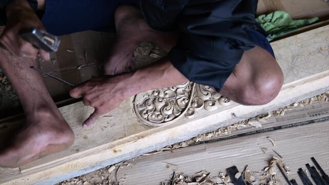 Wood carving work in progress as artisan kneels over ornate board and shapes decorative relief with chisel and mallet. Fresh shavings gather around carved pattern while bare feet brace body