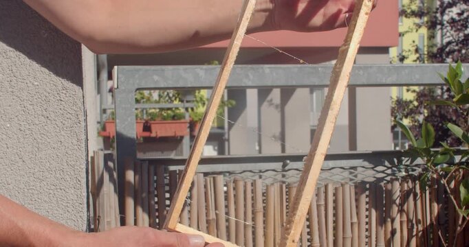 two adult hands lift and inspect wooden beehive comb covered with wax residue and thin wires, tilted during care routine on apartment balcony in sunlight city light