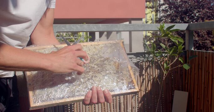 close-up hands peeling clear plastic wrap from wooden tray with coarse granular contents on sunny balcony beside bamboo railing and greenery in natural daylight