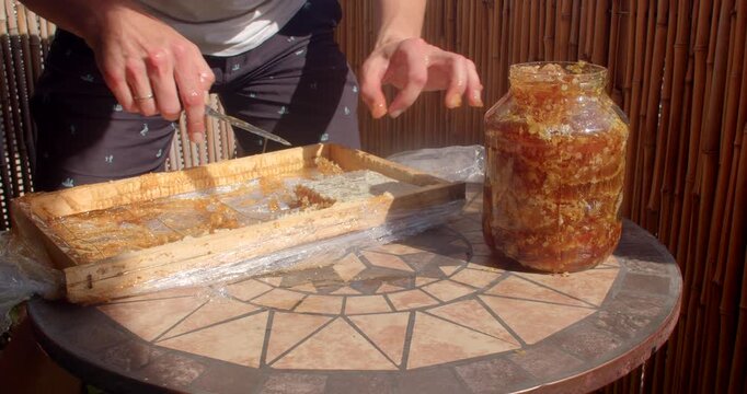 close-up view of hands handling saturated honeycomb over mosaic table while transferring square of comb onto white plate, sticky amber honey drips from comb and