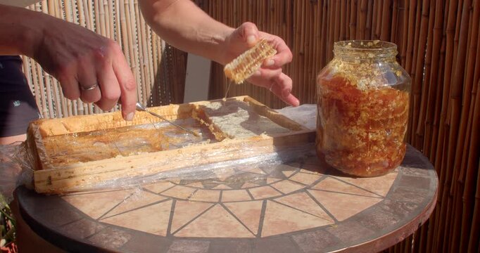 close-up motion as hands lift and separate sticky honeycomb from wooden rack, honey jar filled with amber wax fragments sits on mosaic tabletop, warm sunlight and