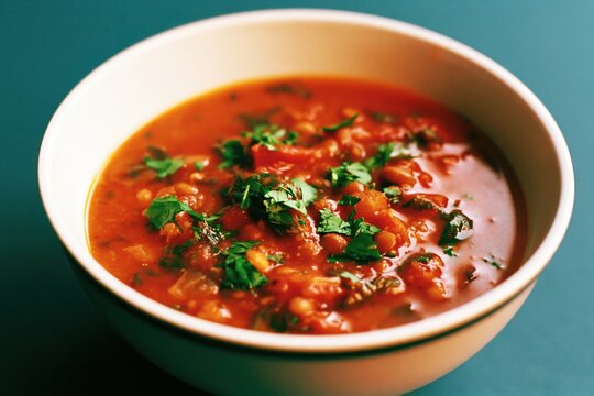 A bowl of harira soup sits on a table with a teal background. The soup is steamy and inviting