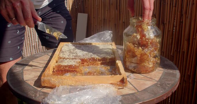 person slices honeycomb and drops amber beeswax pieces into wide-mouth glass jar on small outdoor mosaic tile table, honey dripping in sunlight with close detail