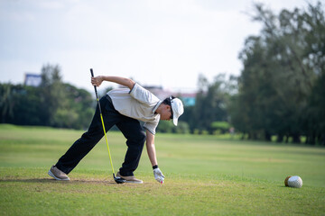 Obraz premium Asian Golfer Placing a Golf Ball on a Tee While Holding a Driver and Preparing for a Focused Tee Shot on a Sunny Outdoor Golf Course