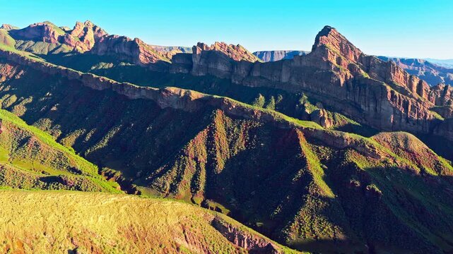 Aerial view of rugged mountain ranges with green ridges, deep shadowed valleys, layered cliffs, and warm sunlight beneath a clear blue sky.