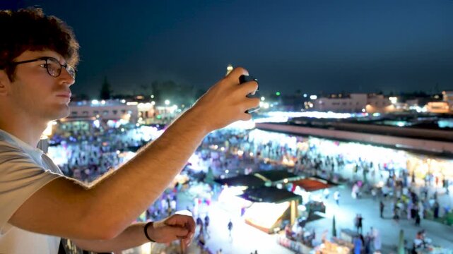 Marrakech, morocco. August 9, 2026. Profile of a young man with glasses observing and taking pictures of the vibrant jemaa el fnaa square at night from a high vantage point
