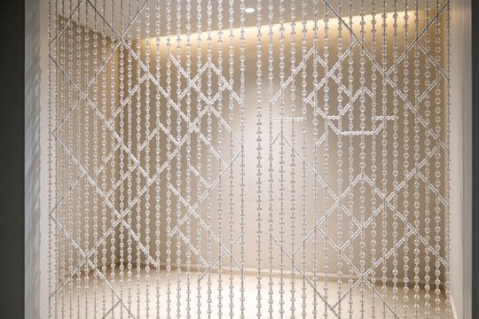 White beaded curtain with geometric pattern hangs indoors. Soft light highlights texture, creating elegant room decor. Translucent material adds subtle privacy and style.