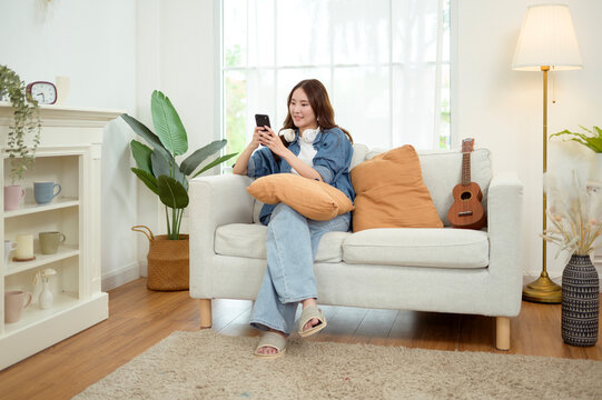 Asian Woman Using a Smartphone While Sitting on a Couch With a Pillow in a Bright Room