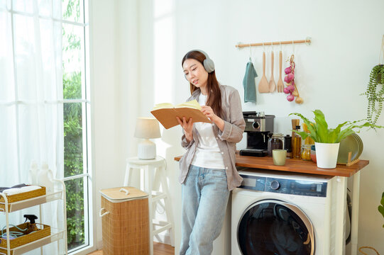Young Woman Reads Book While Standing Next to Washing Machine in Home Kitchen