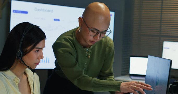  Two Asian female representatives talking at their desk. Professional call center staff reviewing insurance policies, sharing advice, and improving client interaction skills.