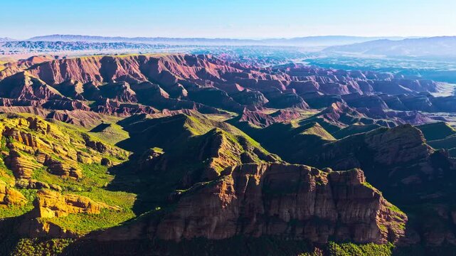 Aerial view of vast eroded mountain valleys and layered red rock ridges, with green vegetation in deep ravines under bright sunlight.