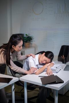 Concerned Asian Businesswoman Waking Up Exhausted Sleeping Colleague at Office Desk Late at Night