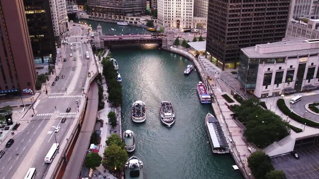 High aerial drone view over the Chicago River with tour boats, bridge traffic, riverfront buildings, and downtown streets during evening light.