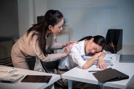 Asian Businesswoman Sleeping at Desk While Colleague Gently Wakes Her During Late Night Office Work