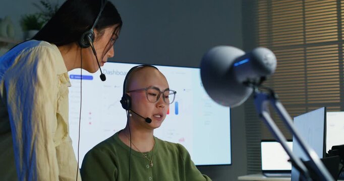 Two Asian women providing digital assistance in a contact center. Dedicated employees wearing headsets, looking at computer screens, and offering reliable technical support.